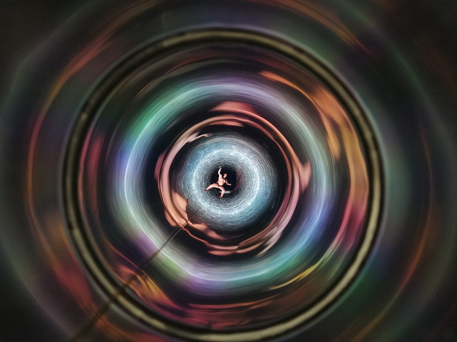Well of Fluid Identities — deep view into the cylindrical interior with iridescent colour reflections creating a vortex-like optical illusion around the figure.