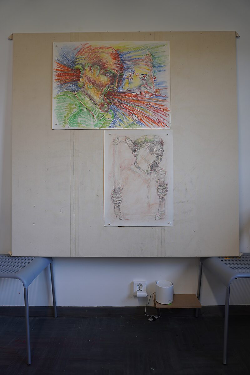 Groot werkpaneel rustend op twee stoelen, met bovenaan een kleurrijke expressieve tekening van een schreeuwende figuur en daaronder een lichtere schets van een figuur die een kader vasthoudt; stopcontact en kleine witte cilinder zichtbaar onder het paneel