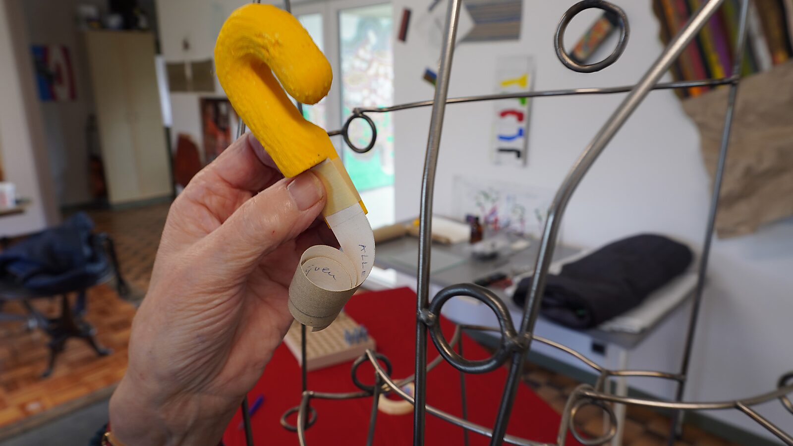 Sfincterman No.0.0 — action videostill: the hand brings the yellow object with the paper strip toward a metal structure with rings in a studio setting.