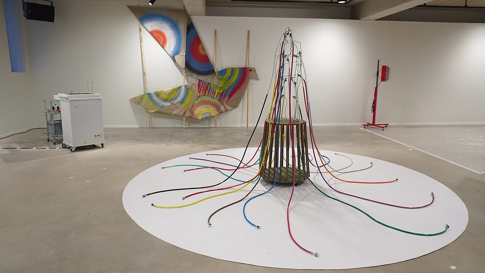 Transformatiestudio nr.1: warm of koud: Tentoonstellingszicht met gevouwen vogelvorm op de achtergrond; beschilderde doeken en een sculptuur met gekleurde buizen op de voorgrond, CC De Adelberg, Lommel, 2024.