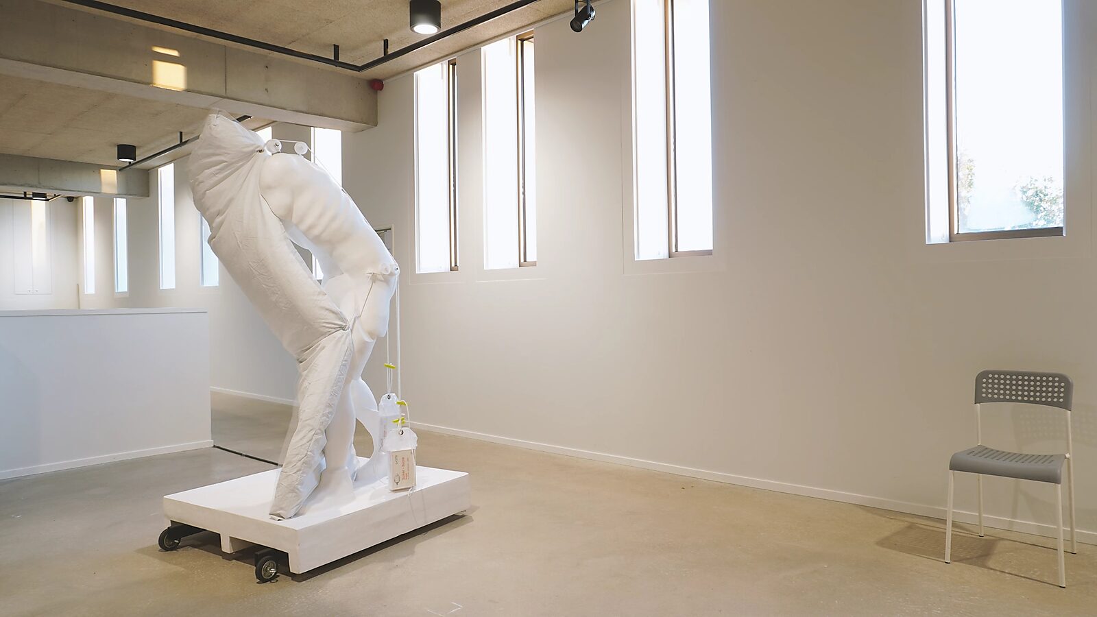 Uitslapen met de slapers — videostill van het kunstwerk op de tentoonstelling Transformatiestudio nr.1: warm of koud, CC De Adelberg Lommel: een wit beeld van een voorovergebogen figuur drukt tegen een groot luchtmatras dat half opgeblazen naast het licha