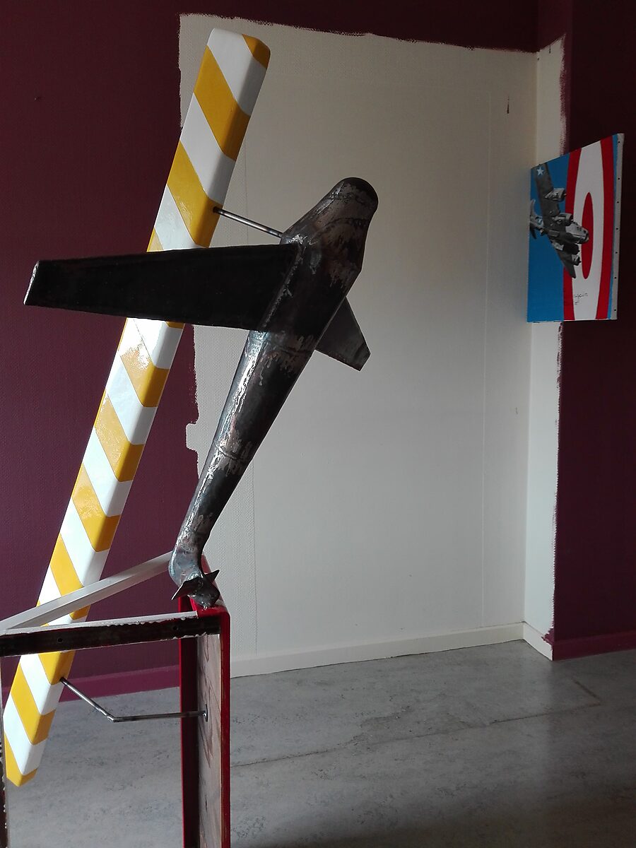 TarmaX: Detail van metalen vliegtuigsculptuur met brede vleugels en zichtbare lasnaden; geel-wit gestreepte balk erachter en klein schilderij met straaljager aan de wand, Tarmac, Meerhout, 2017