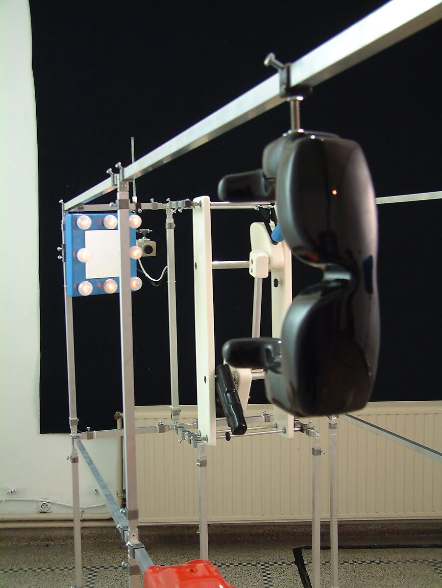 Detail of installation with black sunglasses in the foreground and a metal frame structure with illuminated mirror and mechanical components in the background; Villa Basta, Houthalen.
