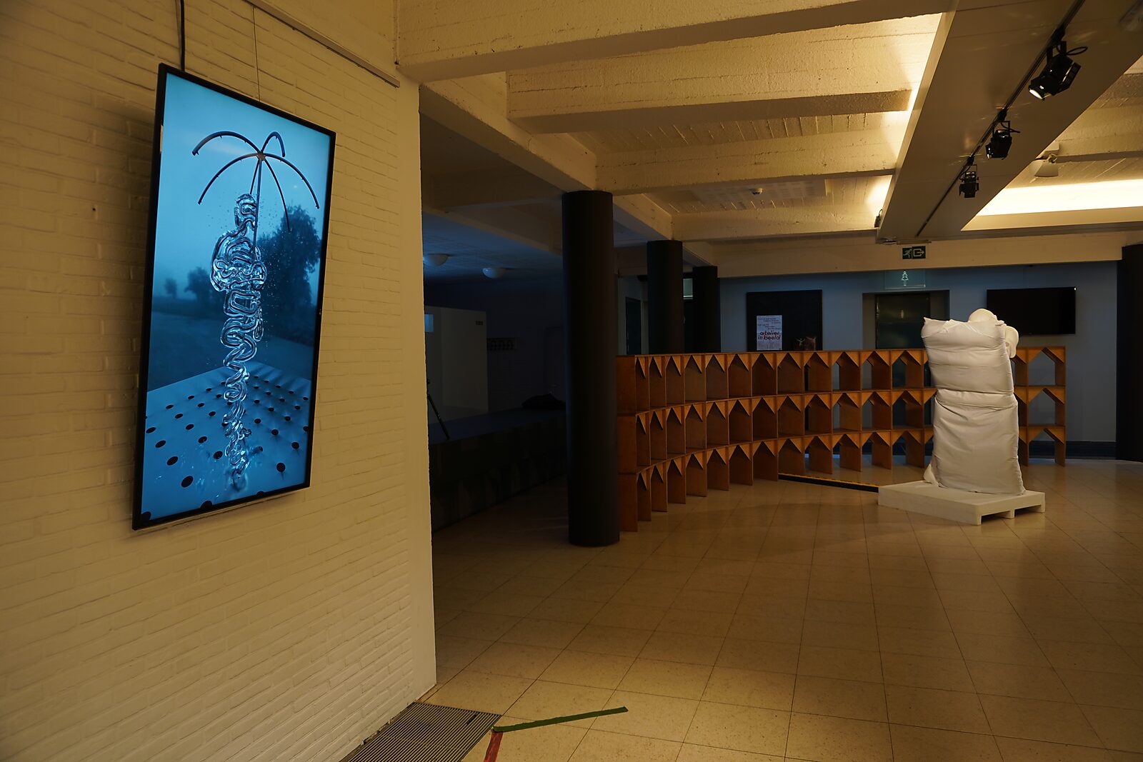 'Sfincterman for President’: Tentoonstellingszicht met verticaal videoscherm (blauw beeld van een sculptuur) en op de achtergrond een witte sculptuur bij een houten structuur, Dommelhof, Pelt, 2024.