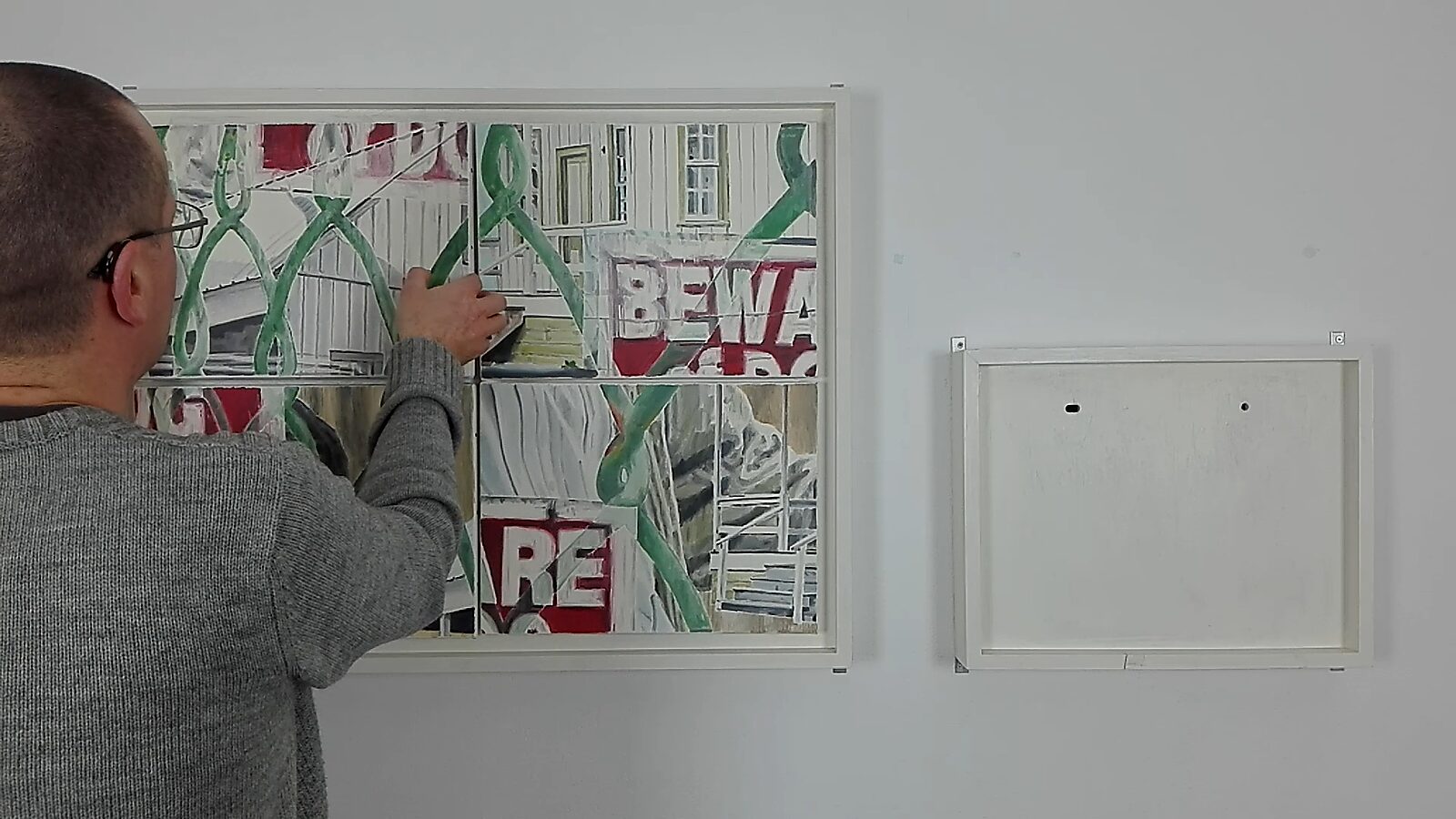 Opgepast voor hond — videostill handeling: een man herschikt panelen binnen een wit kader aan de muur, waardoor fragmenten van een schilderij met groene hekken en rode tekst verschuiven.