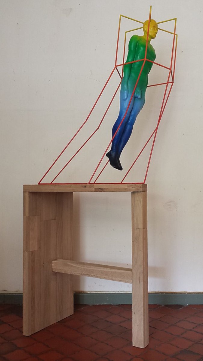 Gedachtenkooi — eerste versie van de sculptuur waarin een menselijk lichaam gevangen lijkt in een geometrisch metalen kader, geplaatst op een houten sokkel en uitgevoerd in kleurverlopen die verwijzen naar warmtecamera-beelden.