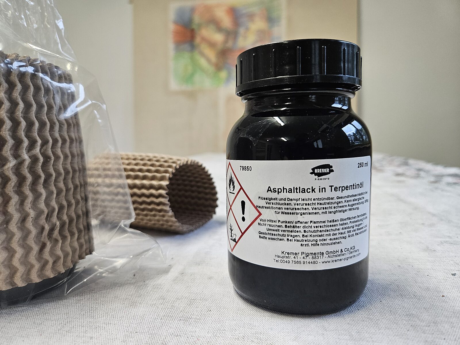 Black jar labeled “Asphaltlack in Terpentinöl” next to protective packaging material on a table; in the background, a drawing mounted on a panel appears out of focus.