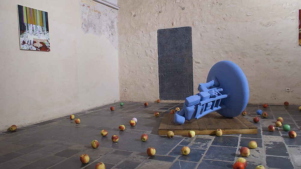 ‘Strange Fruit’: Blue robot figure fallen over on a platform with a circular disc; real apples scattered across the floor, painting on the wall, Gasthuiskapel, Borgloon, 2015.