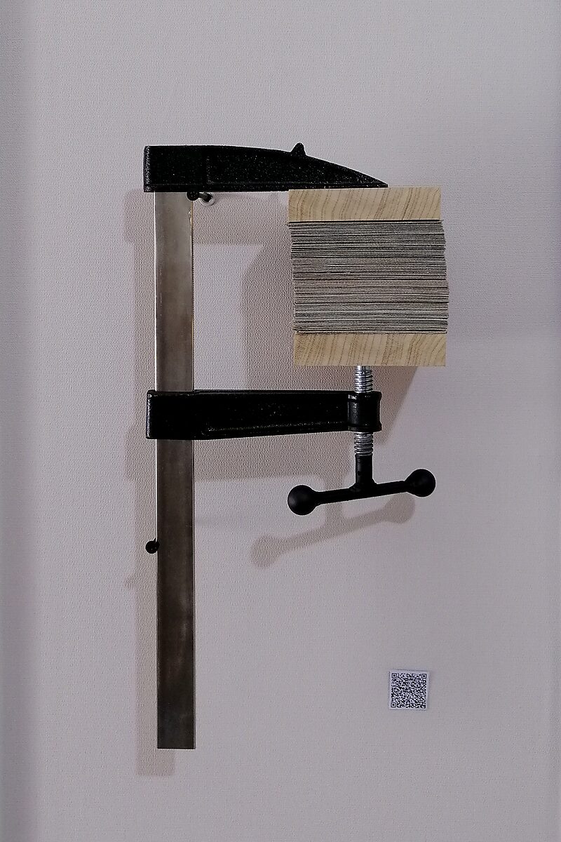 Paintings.zip — a cut painting is compressed between wooden blocks in a metal clamp mounted to the wall; the pressure of the clamp compresses the stacked layers of canvas and paint into a compact block.