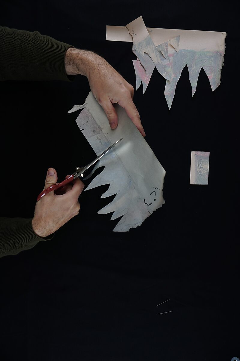 Benoemens-waardvlinders — Naming–Value Butterflies — proces tekening: Robotman (1995) — handen knippen een klein stukje papier met een naamfragment uit terwijl de resten van de tekening erboven hangen.