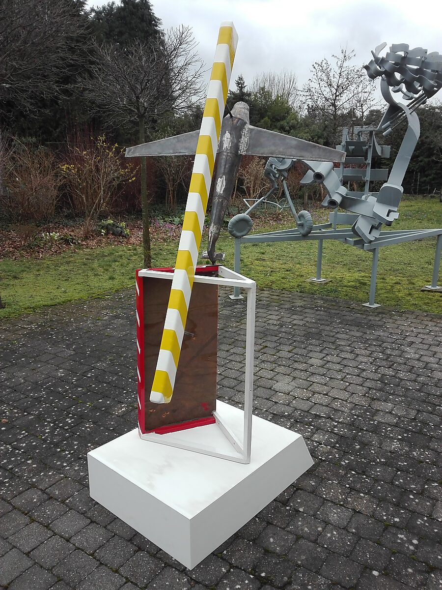 Gelovige vliegtuigen — sculptuur met metalen vliegtuigvorm die schuin balanceert op een geometrisch frame met rood-witte en geel-witte diagonale strepen, opgesteld op een witte sokkel in een buitenomgeving met andere sculpturen op de achtergrond.