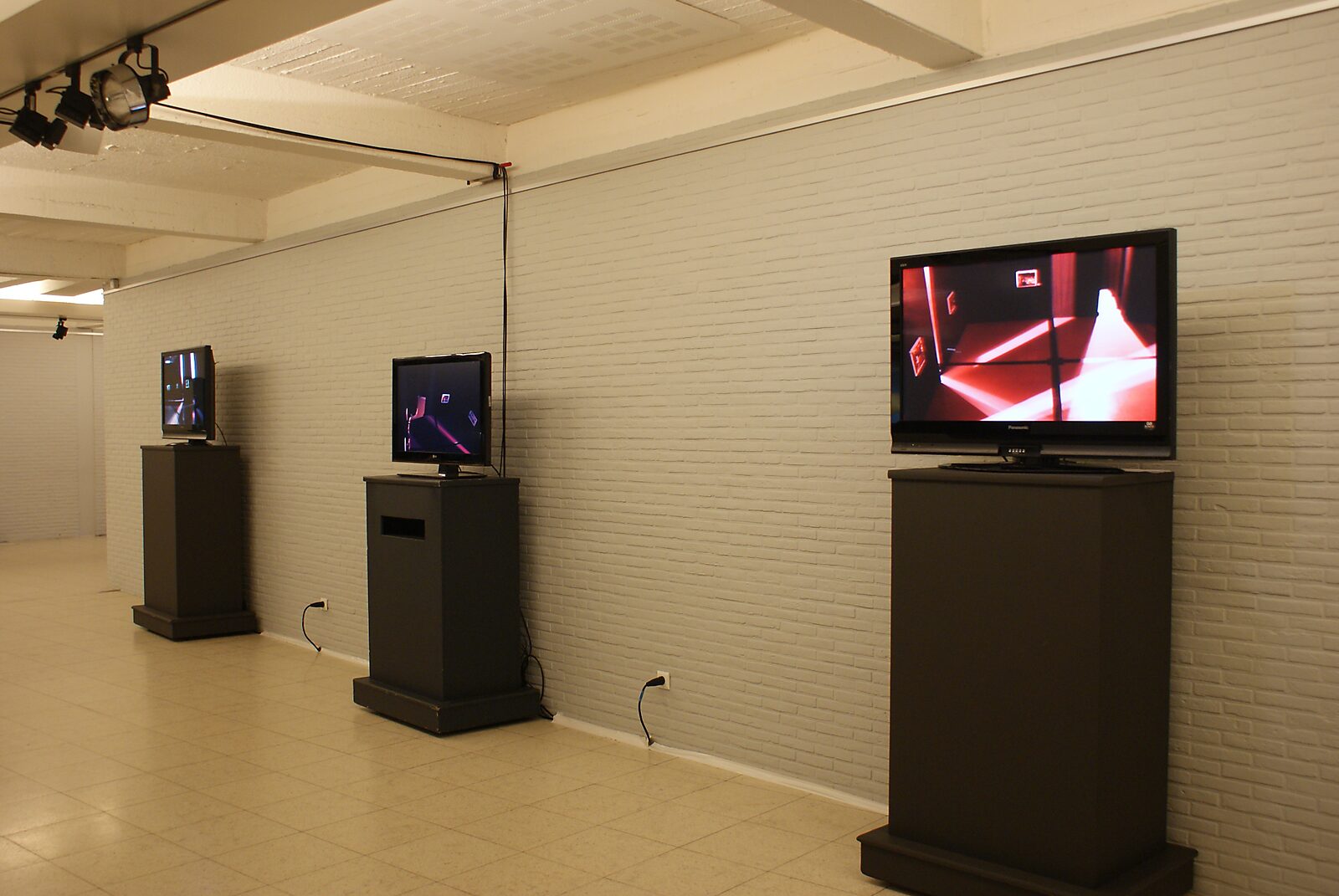 The Modern Cavemen — exhibition space at C-TAKT Pelt, Dommelhof with three television monitors on pedestals; the screens display camera views of the exhibition itself, turning the space into something simultaneously observed and mediated.