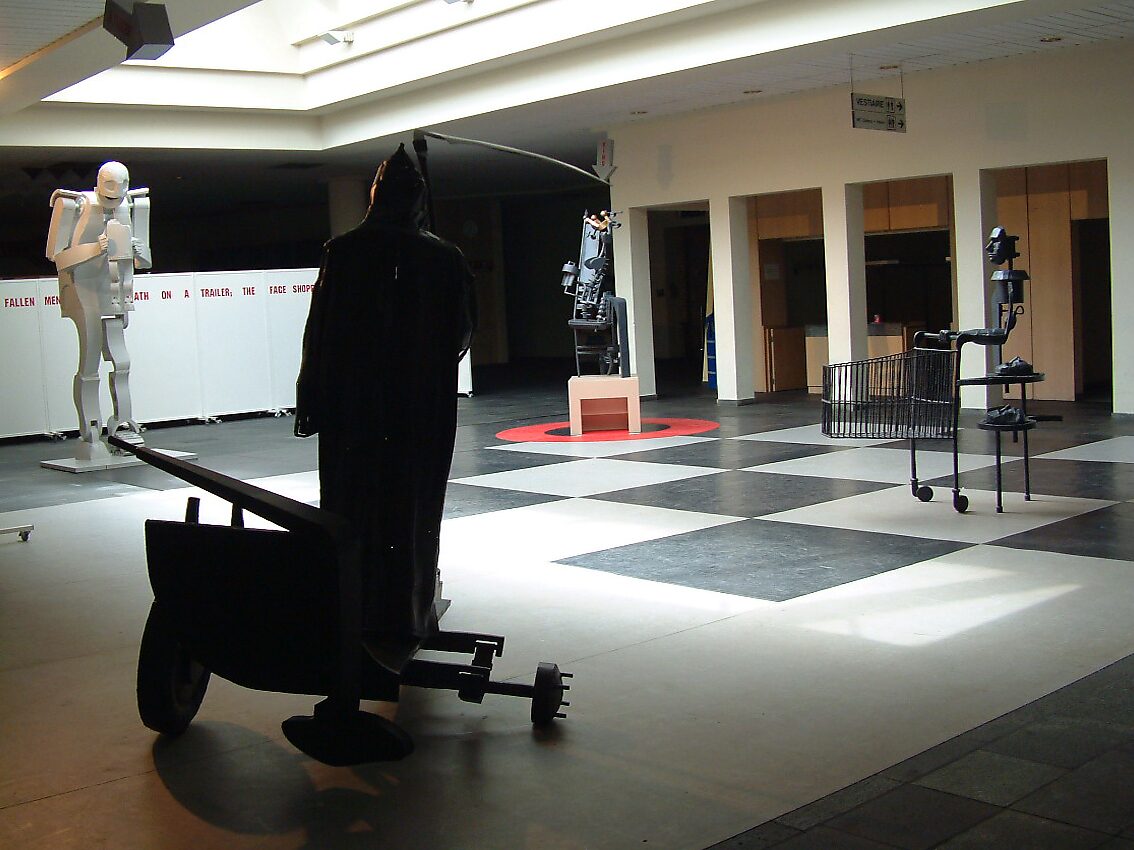 ‘Ode to Saturn’: Tentoonstellingszicht met meerdere metalen figuren in gelast plaatstaal; zwarte figuur met kar op de voorgrond en witte monumentale figuur achteraan, CC De Adelberg, Lommel, 2003.