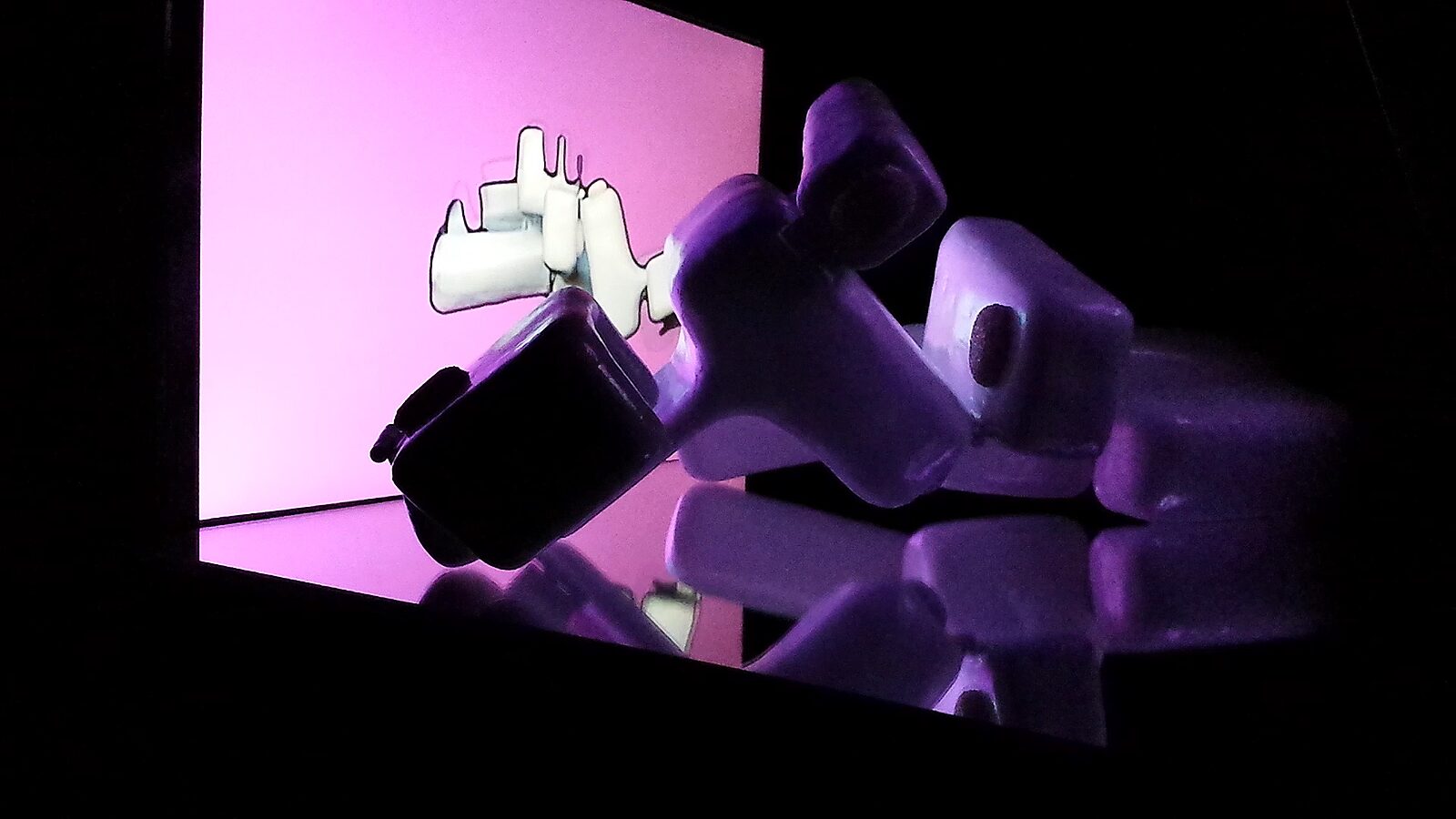 Reflecties in beweging — videostill opname: het beeld kantelt en reflecteert in het donkere oppervlak, terwijl het scherm achteraan de vorm vervormd en fragmentarisch weergeeft.