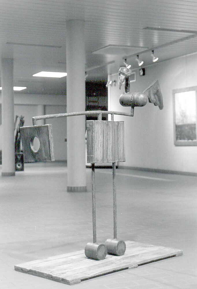 ‘Houten spiralen en metalen’: Metalen vogelachtige figuur met houten elementen op rechthoekige sokkel; sculptuur in hout en metaal, zwart-wit tentoonstelling, CC Leopoldsburg, 1999.
