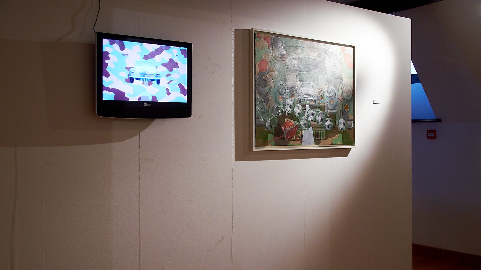 Duo exhibition with Raymond Minnen: Exhibition view with wall-mounted monitor showing a digital animation of a car in camouflage colours; next to it a large painting depicting a frontal view of a vehicle, CC De Kimpel, Alden Biesen, 2011