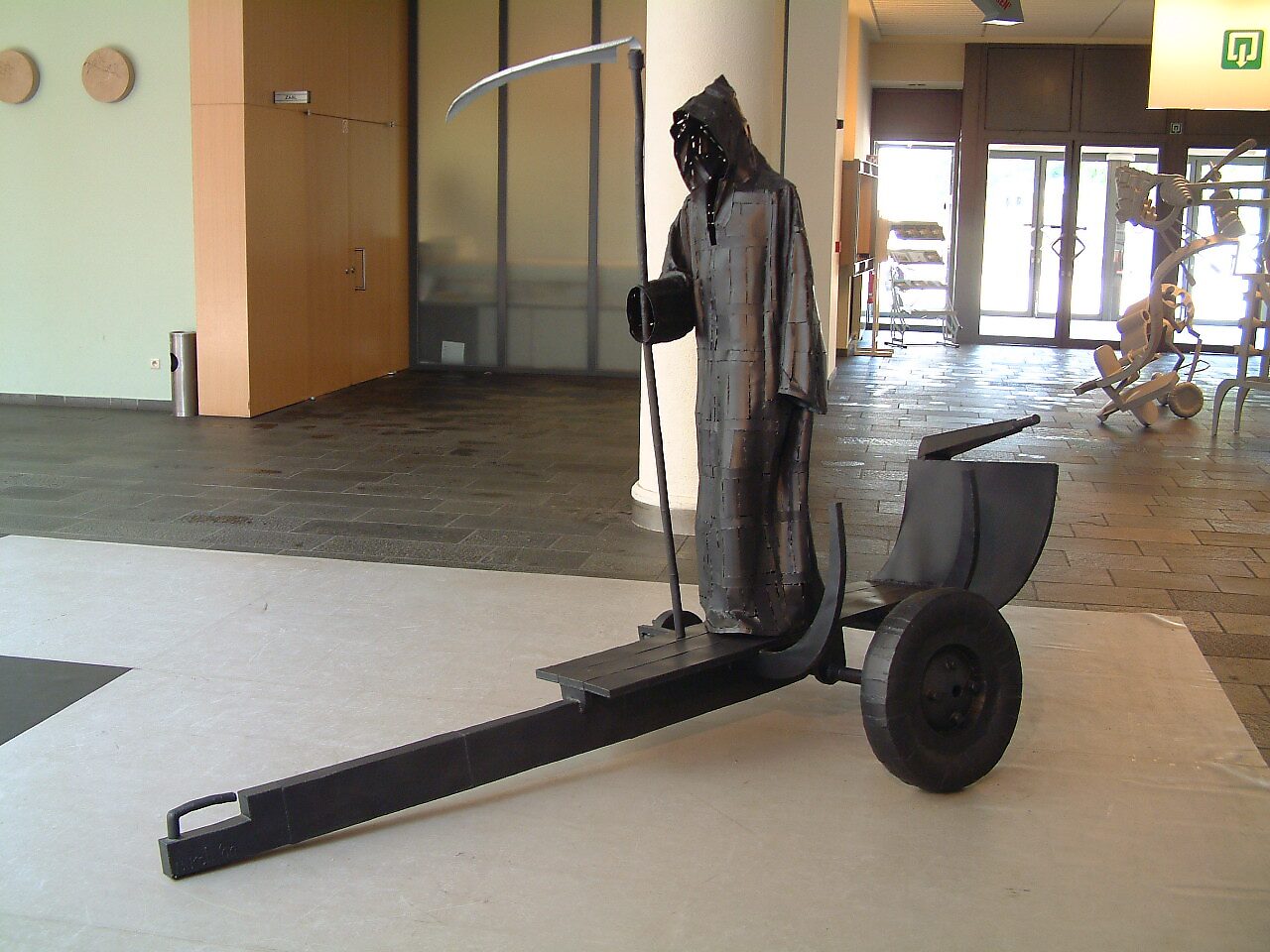 Onderbroken overtocht — zwarte metalen sculptuur van Pietje de Dood als veerman met zeis, staand op een boottrailer in tentoonstellingsruimte CC. De Adelberg.