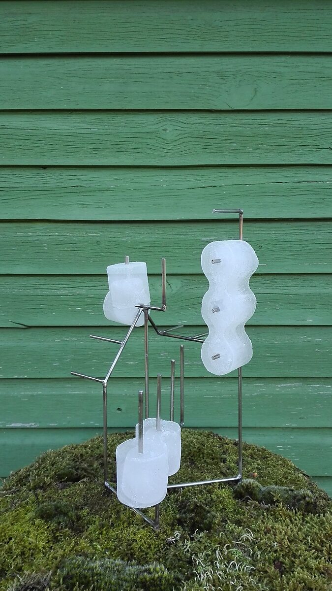 Entropische sculptuur nr. 1 — videostill van een metalen armatuur met ijsvormen erop bevestigd, geplaatst op een met mos begroeide heuvel voor een groene houten wand, opgenomen in de lente.