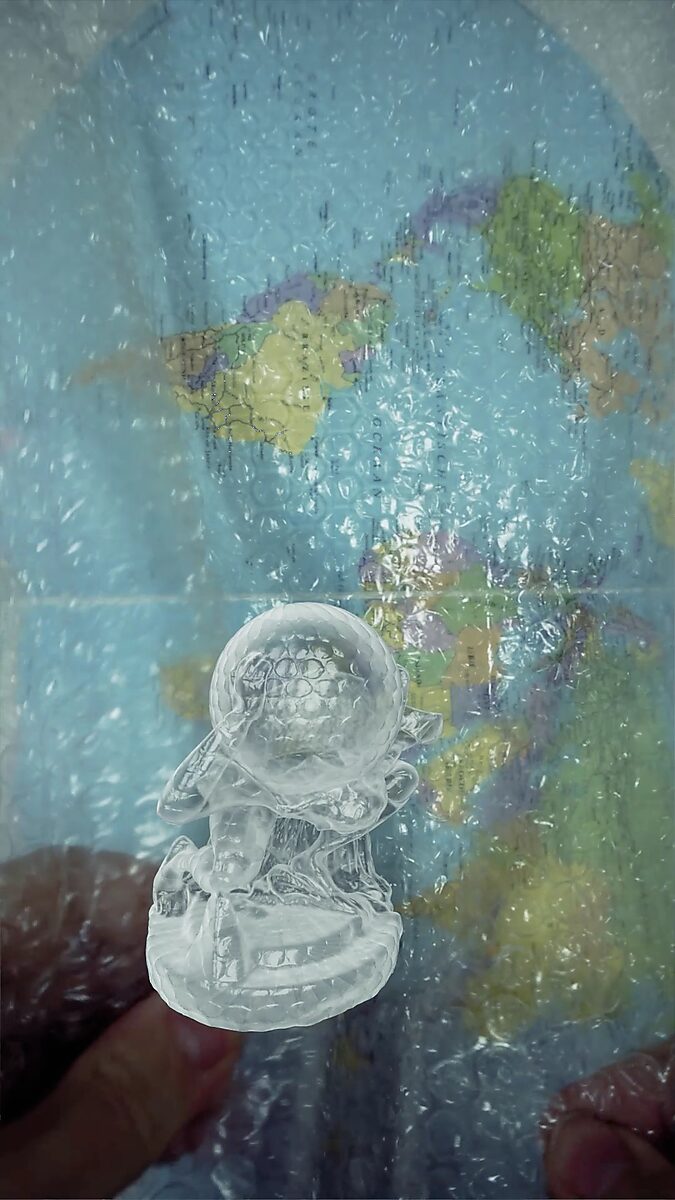 Virtuele transformatie — Atlas in bubbelplastic: videostill uit een 3D-animatie; de figuur zakt licht door zijn knieën terwijl de bol vervormt en het bubbelplastic de kaart erachter vervaagt en vervormt.