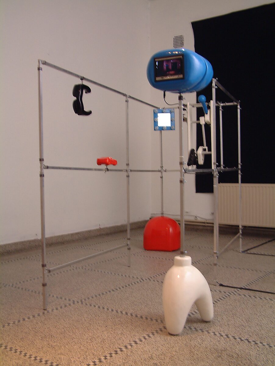 Kies en verander, Installatie voor een egoïstische rapper — foto van de installatie: modulaire metalen structuur met hangende objecten, waaronder een blauw monitorachtig element, zwarte en rode vormen en een witte sculptuur op de vloer.