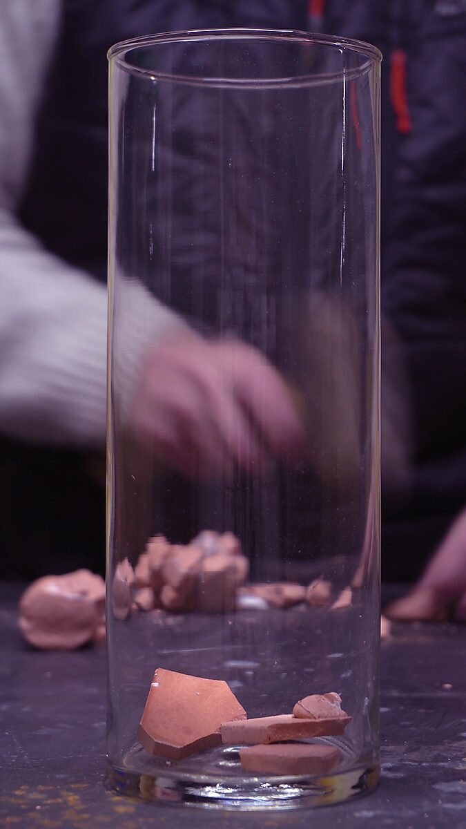 Gebroken volume — videostill met verspreide terracotta fragmenten in de glazen cilinder.