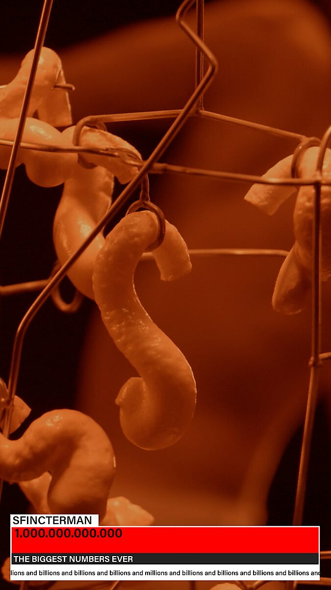 Sfincterman — videostill videowerk: oranje belichting toont een close-up van een hangende vorm, met op de achtergrond een onscherp beeld van een naakte danser.
