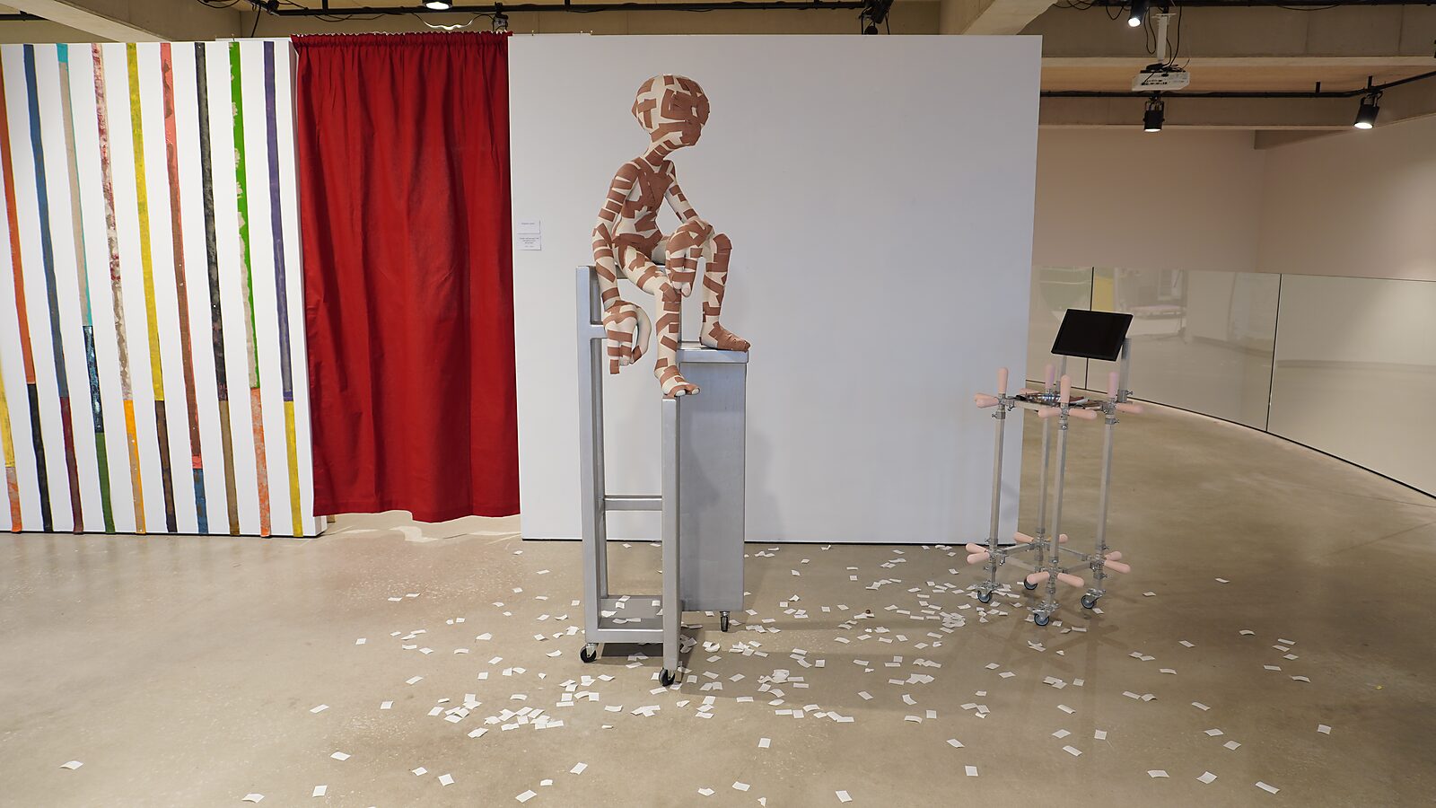 Buitenaardse zorg, pleisterfase 1 — ruimtelijk zicht: installatie met alienfiguur op hoge stoel, rode gordijnpartij, verticale schilderijen en metalen constructie; papiersnippers verspreid over de vloer.