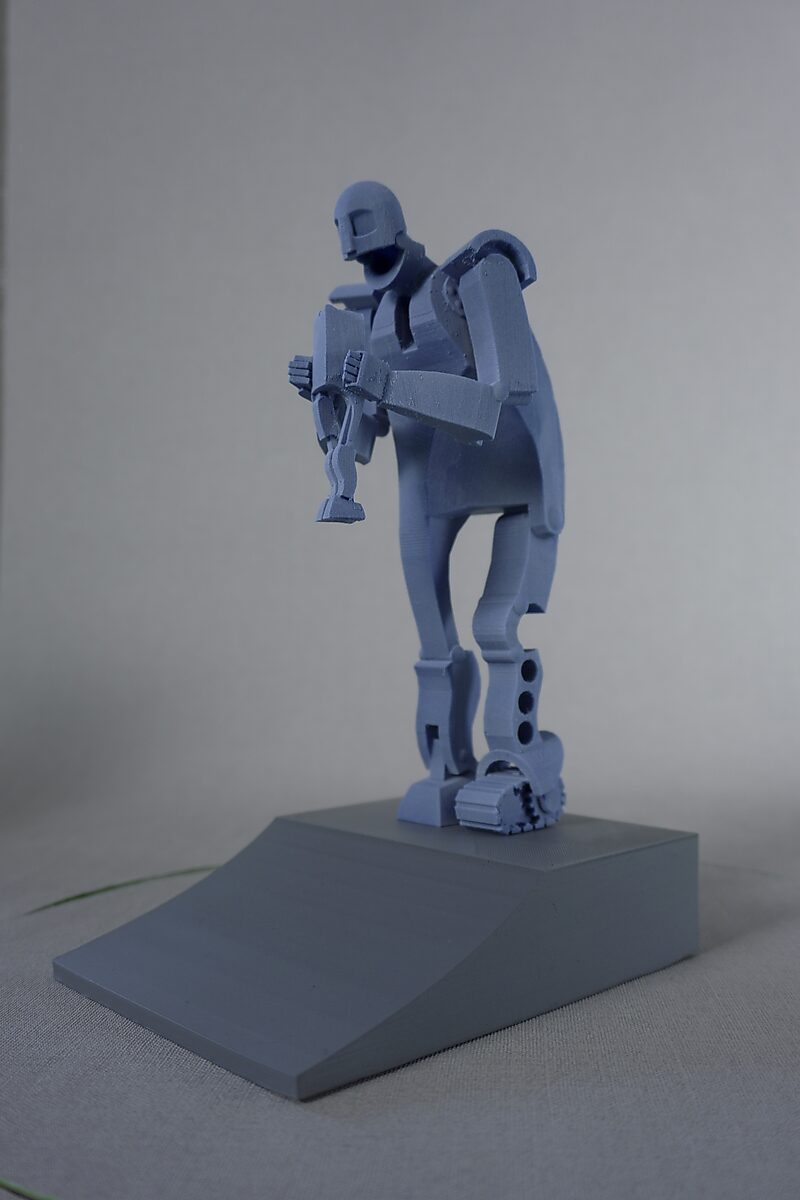 Saturn — blue 3D-printed miniature of a standing figure holding a smaller figure, displayed on a grey inclined pedestal.