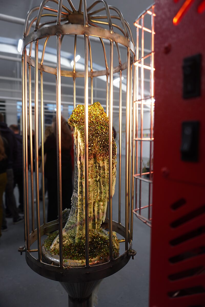‘Malabou in Malibu’: Detail of cage containing a glittering, partially melted figure; warm lighting and red installation element in the background, De Fabriek, Eindhoven, 2023.