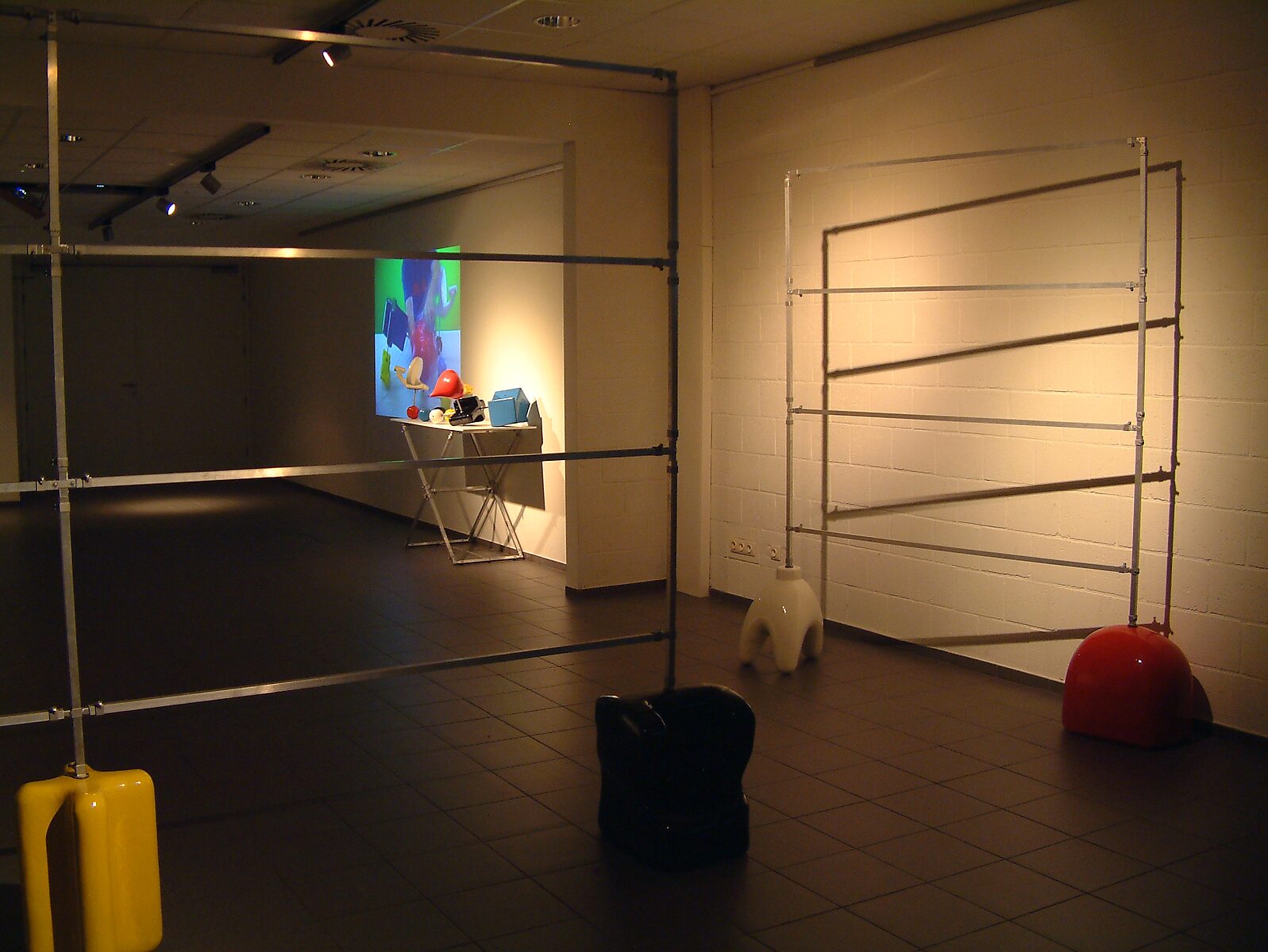 'Pick and Choose Project’: Exhibition view with metal tube construction against a white wall; video projection and table with objects visible in the adjacent space, CC De Kollebloem, Puurs, 2008.