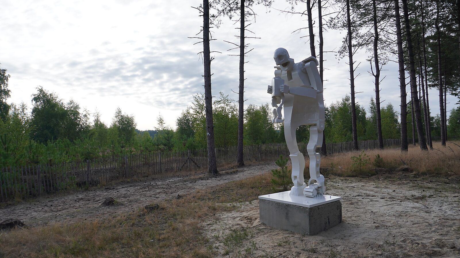 Saturn — overall view of the white sculpture within its environment, the figure standing centrally in the sandy terrain of the Sahara in Lommel, contrasting with the surrounding forest and open sky.