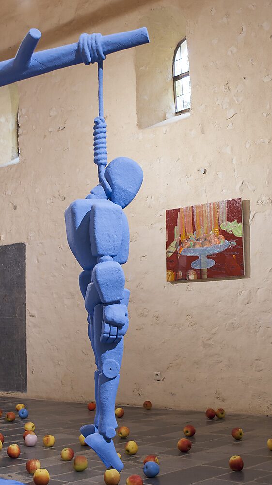 ‘Strange Fruit’: Detail of a blue robot figure hanging from a beam; real apples on the floor and a painting in the background, Gasthuiskapel, Borgloon, 2015.