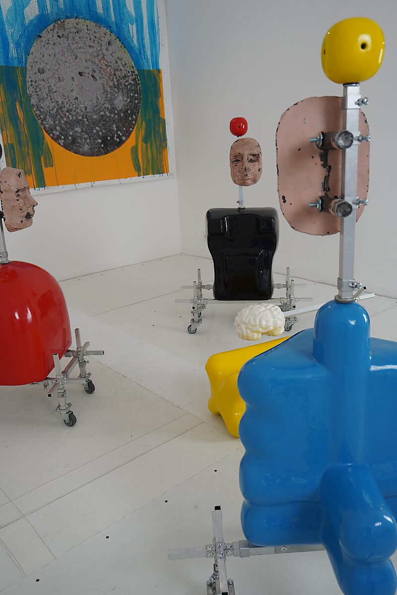 Group of colorful sculptural elements on wheels, including stylized heads and torsos in red, blue, and black, arranged in a white space with a painting in the background.