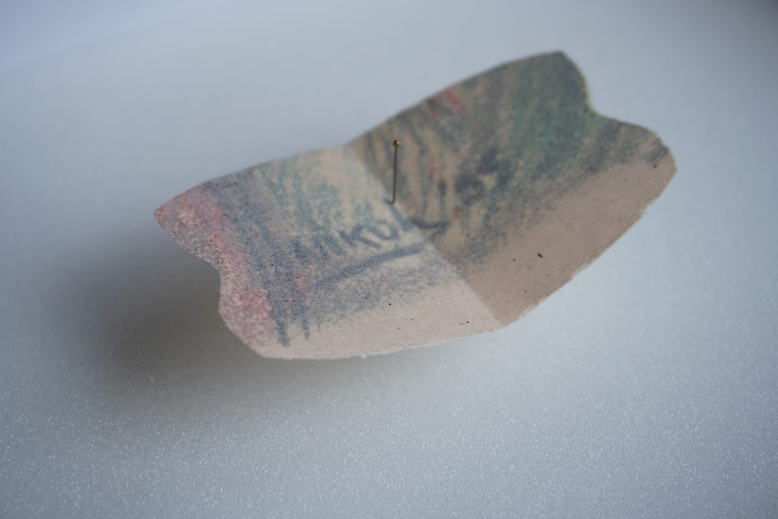 Naming–Value Butterflies — process drawing: Hanging Man (1995) — a small irregular piece of thin paper with faint color traces lies on a white surface; a thin pin stands upright in the center.