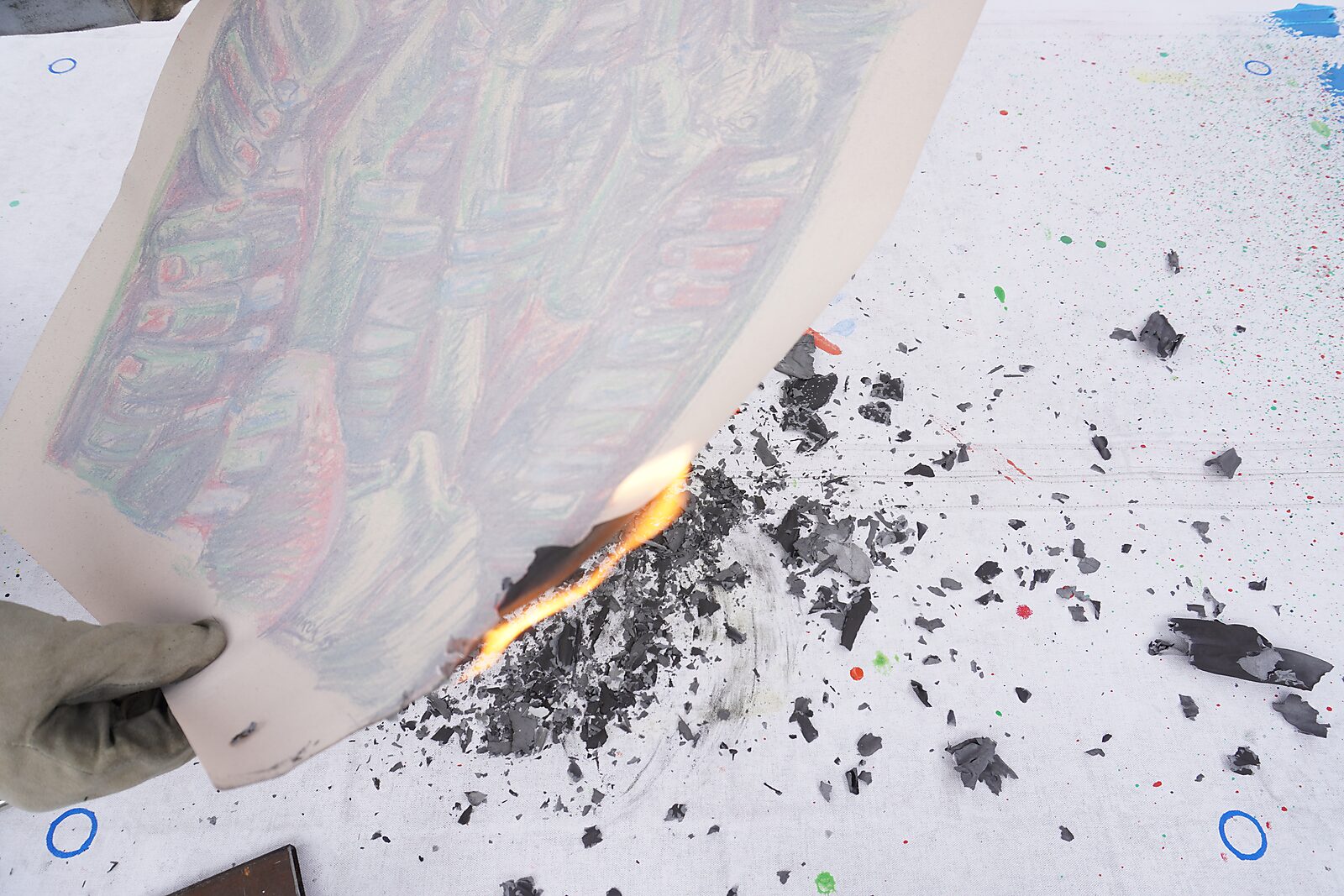Naming–Value Butterflies — process drawing: Hanging Man (1995) — a hand holds the drawing while the edge of the paper burns; black ash and charred fragments lie on a white table.