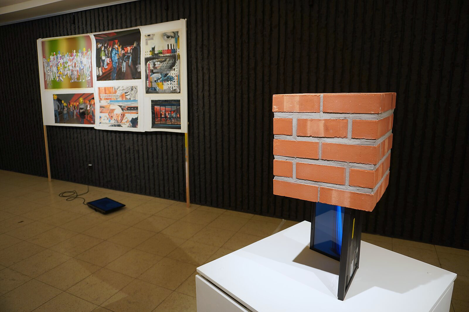 The Digital Age — exhibition view of the sculpture displayed on a pedestal, showing the opened laptop and brick structure, with other artworks and projections visible in the background.