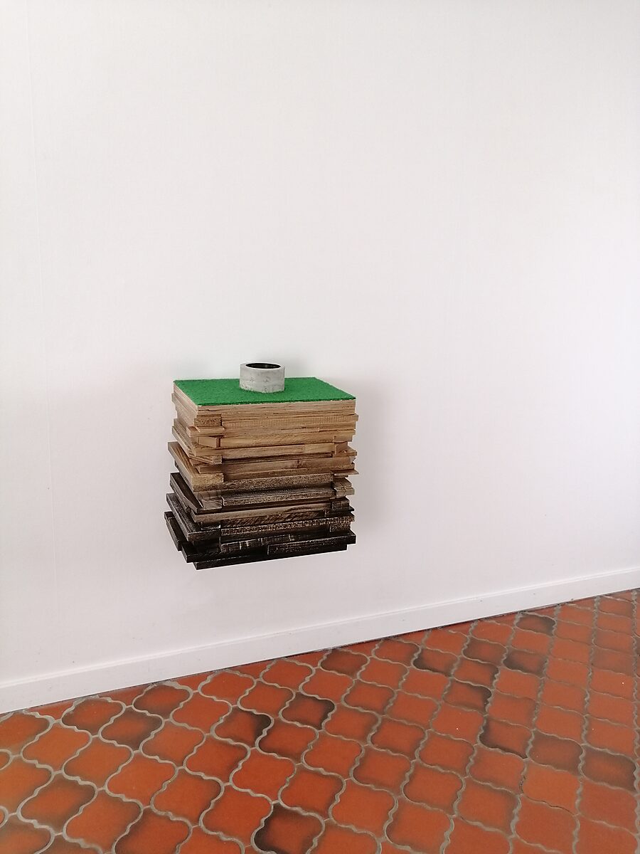 Well of Fluid Identities — wall-mounted sculpture composed of stacked wooden boards, topped with green artificial turf and a circular concrete well opening, with red tiled floor visible below.