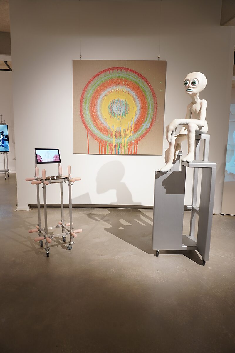 Buitenaardse zorg, pleisterfase 1 — tentoonstellingszicht: alienfiguur zit op een hoge grijze stoelconstructie naast een schilderij met kleurrijke concentrische cirkels; in de ruimte staat een metalen installatie met roze elementen.