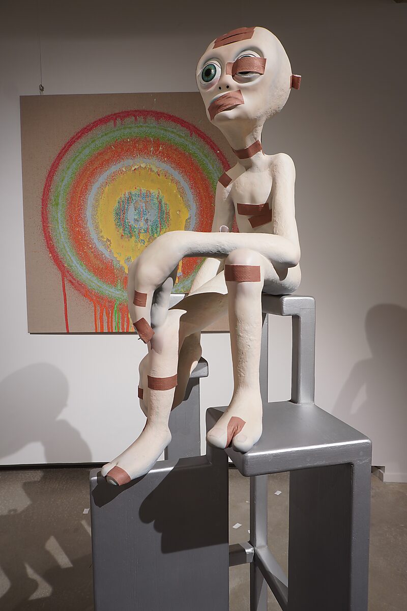 Buitenaardse zorg, pleisterfase 1 — detail: alienfiguur zittend op een hoge stoel, gedeeltelijk bedekt met bruine pleisters op gezicht, armen en benen; schilderij met kleurrijke cirkels op de achtergrond.