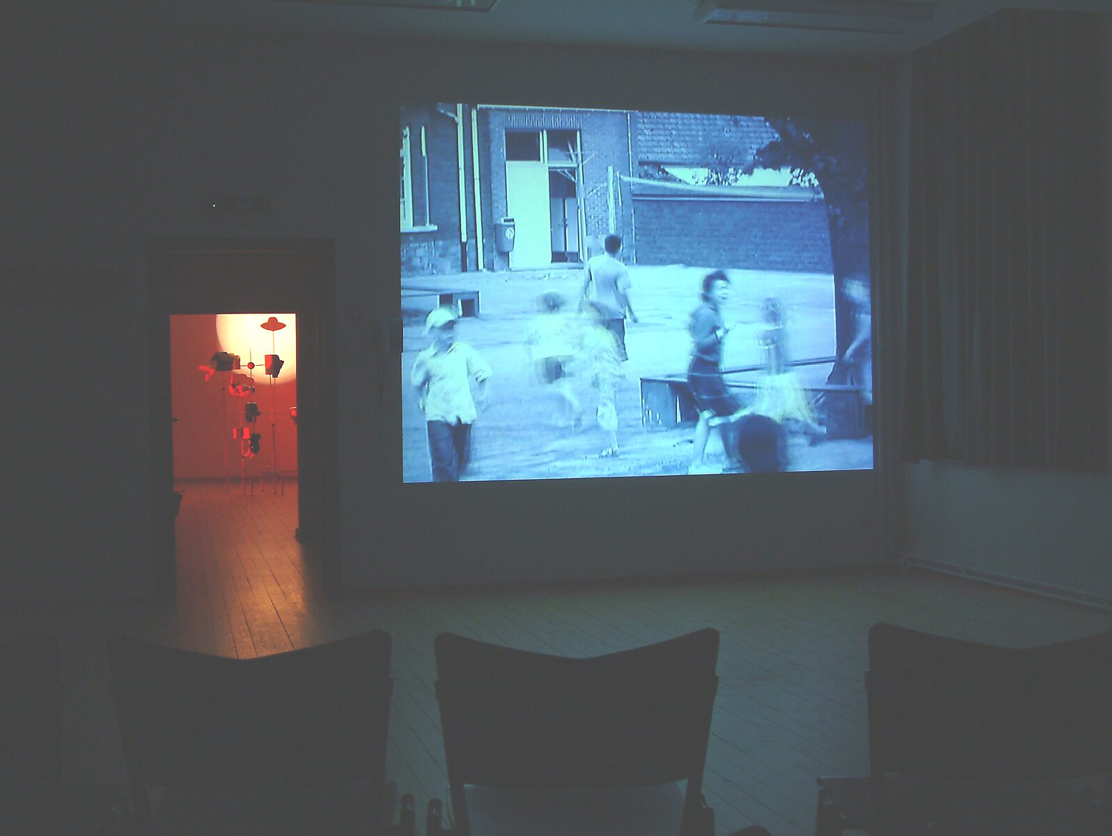 ‘Zien’: Tentoonstellingszicht in verduisterde ruimte met grote zwart-wit filmprojectie van spelende kinderen; links in een aangrenzende ruimte een verlichte sculpturale installatie zichtbaar, CC De Breughel, Bree, 2006.
