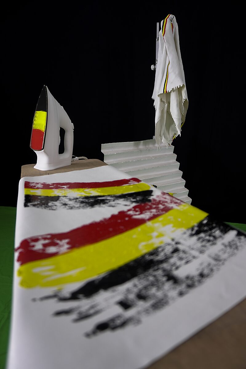 Belgium Smooths Out the Wrinkles — detail view of the cloth hanging over the ironing board while the iron and tricolour paint strokes create a painterly surface between domestic object and painting.