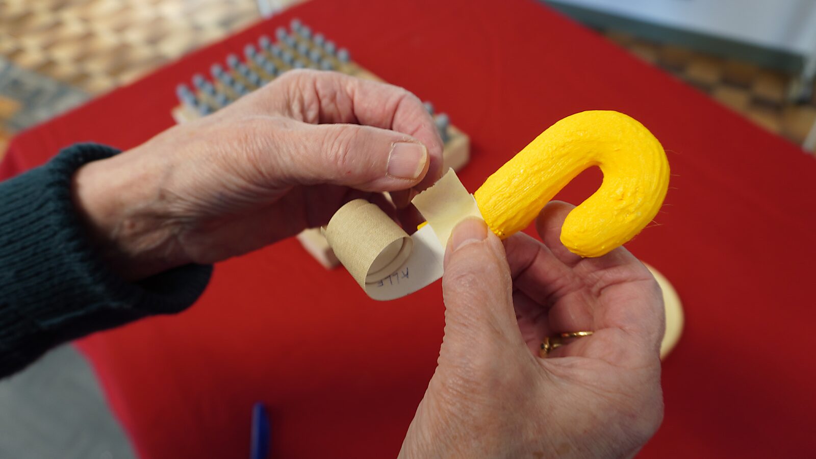 Sfincterman No.0.0 — action videostill: a paper strip is attached with tape to a small yellow hook-shaped object held in the hand.