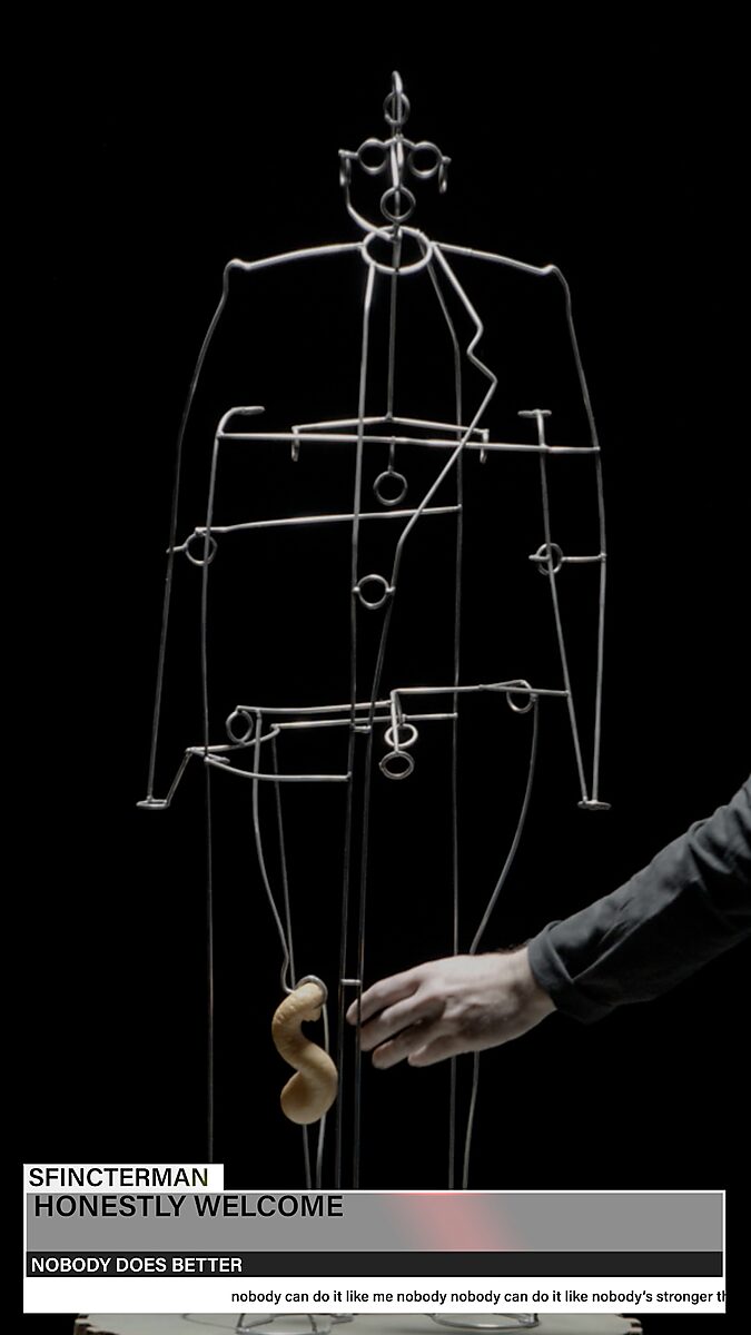 Sfincterman — video work videostill: a metal wire figure stands against a black background while a hand attaches a foam-like form to the body; graphic text bars appear at the bottom.