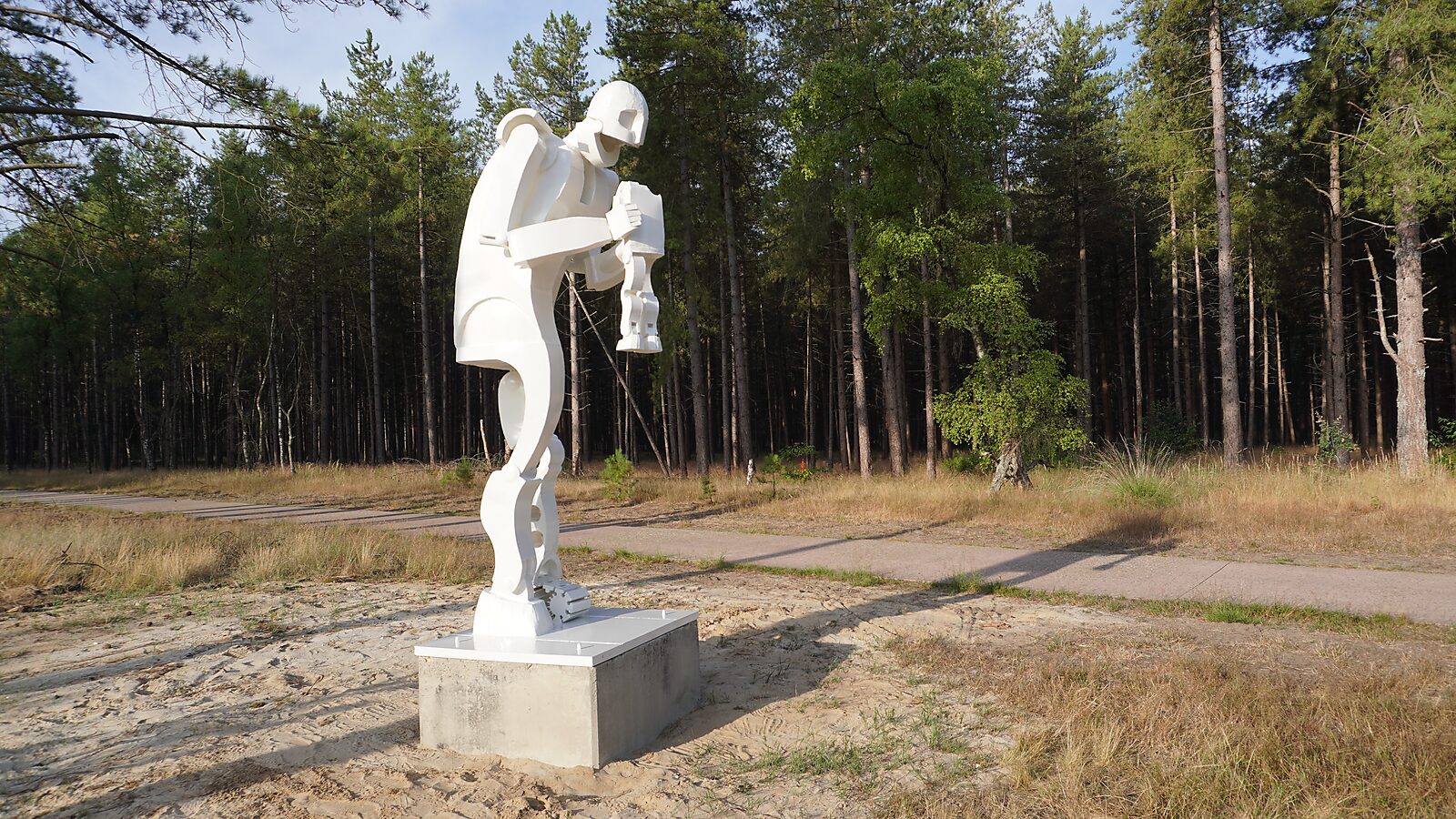Saturn — side view of a white figurative sculpture with mechanically constructed limbs, installed on a concrete pedestal along a forest path in the public landscape of the Sahara in Lommel.
