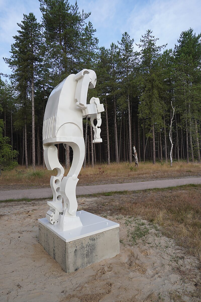 Saturnus — achteraanzicht van een witte sculptuur met gebogen rug en opengewerkte benen, waarbij de figuur een kleiner lichaam naar beneden houdt, geplaatst in een open zandvlakte met dennenbomen.