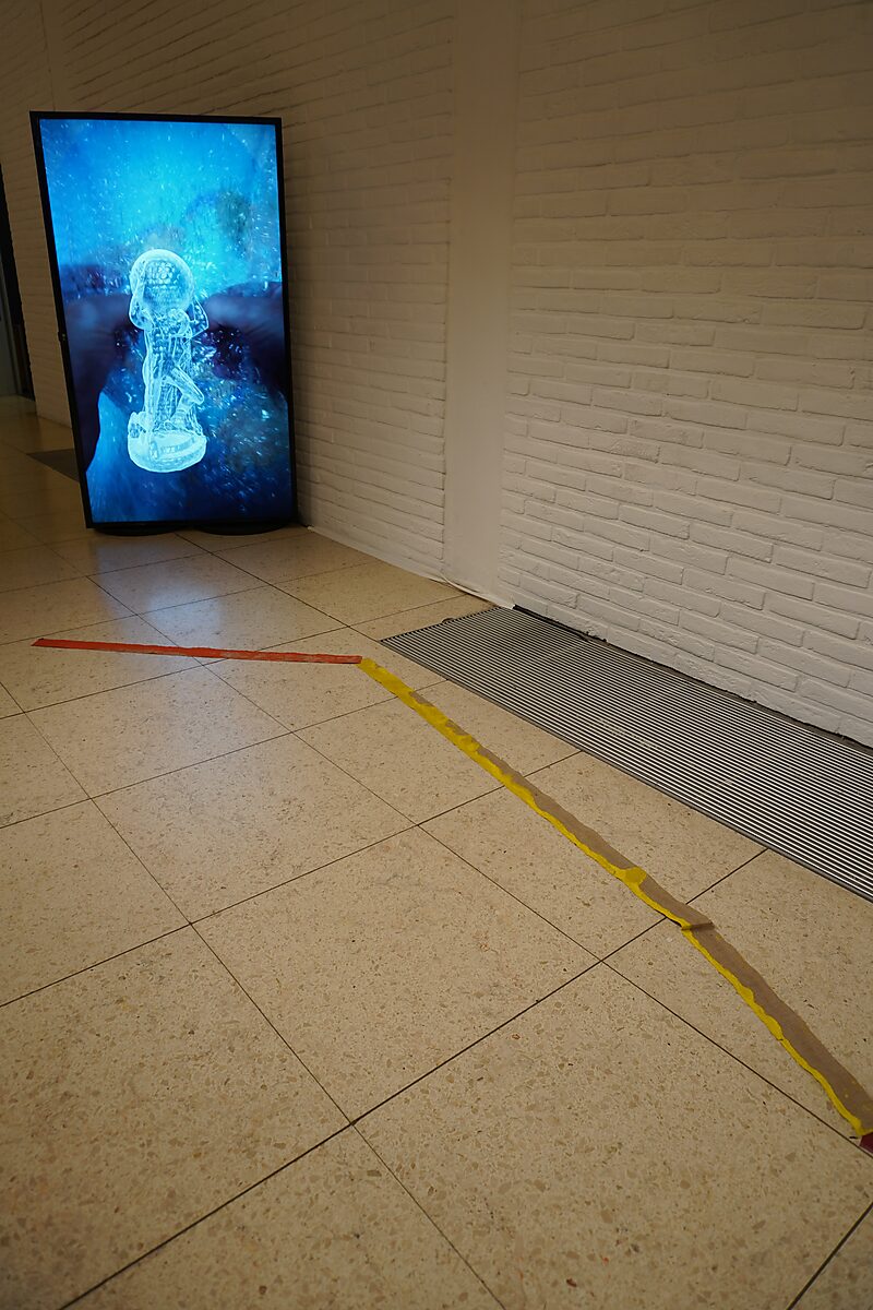 'Sfincterman for President’: Exhibition view with a vertical video screen in a corner; a red-and-yellow line runs across the floor toward the screen, Dommelhof, Pelt, 2024.