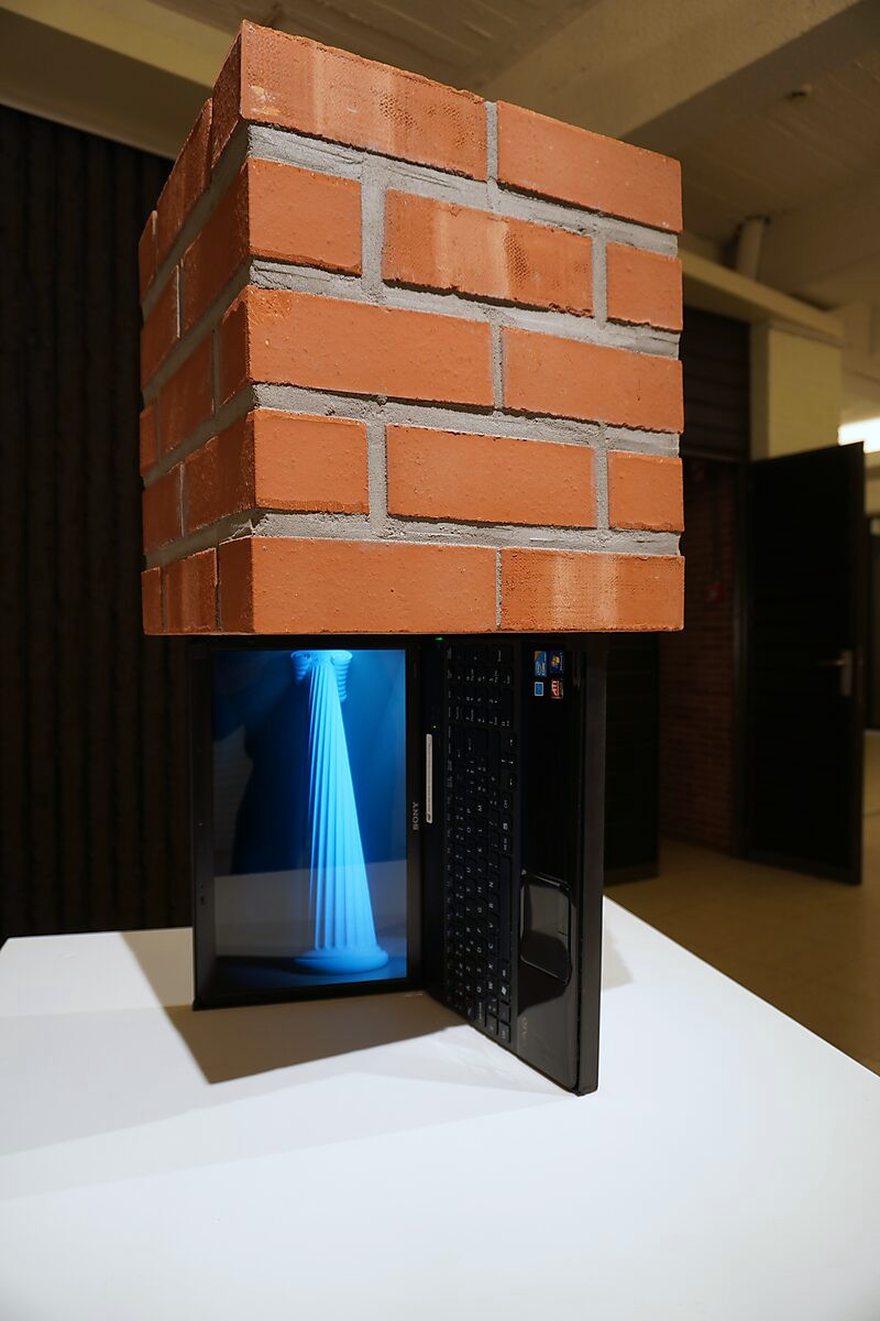 The Digital Age — sculpture consisting of an opened laptop supporting a brick volume; a blue 3D-animated classical column appears on the screen.