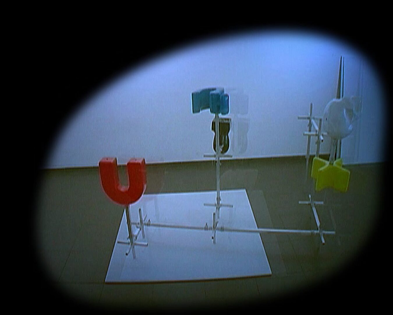 Kies en verander, Installatie in beweging — videostill: metalen constructie met gekleurde vormen (rood, geel, blauw en wit) opgesteld op een witte plaat, gezien door een cirkelvormig kader.