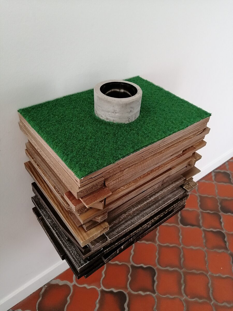 Well of Fluid Identities — angled top view of the layered wooden construction with green surface and a central cylindrical concrete rim marking the well opening.