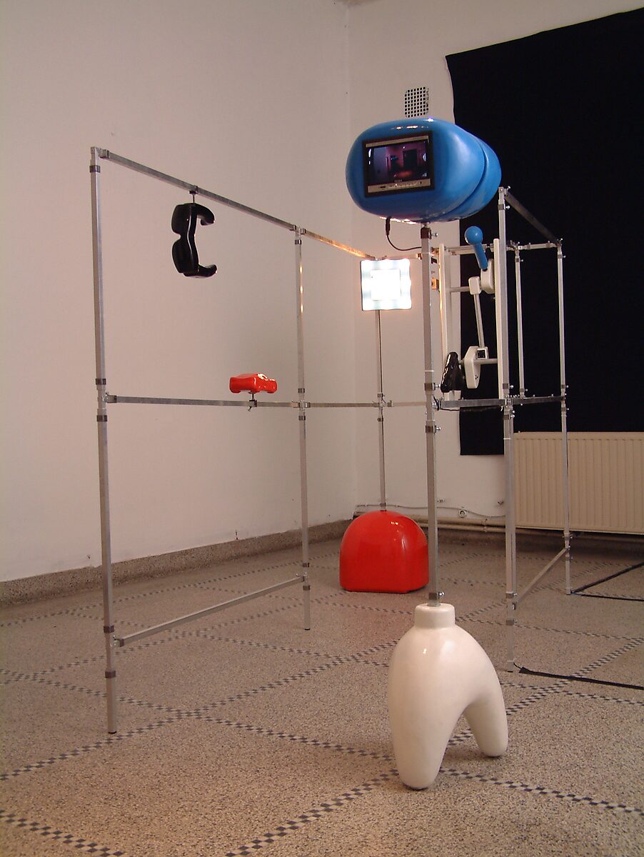 Exhibition view of open metal frame installation with red and white plastic forms on the floor and a blue monitor mounted at the top of the structure; Villa Basta, Houthalen.
